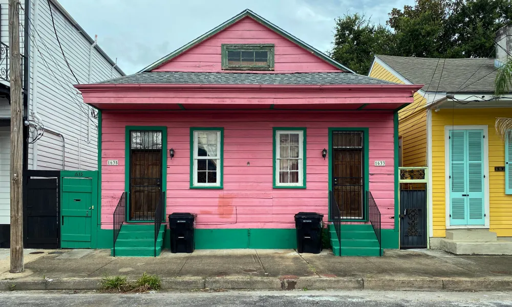 Pink wooden house with green trim, two doors, windows, and trash bins on a city sidewalk in daytime.