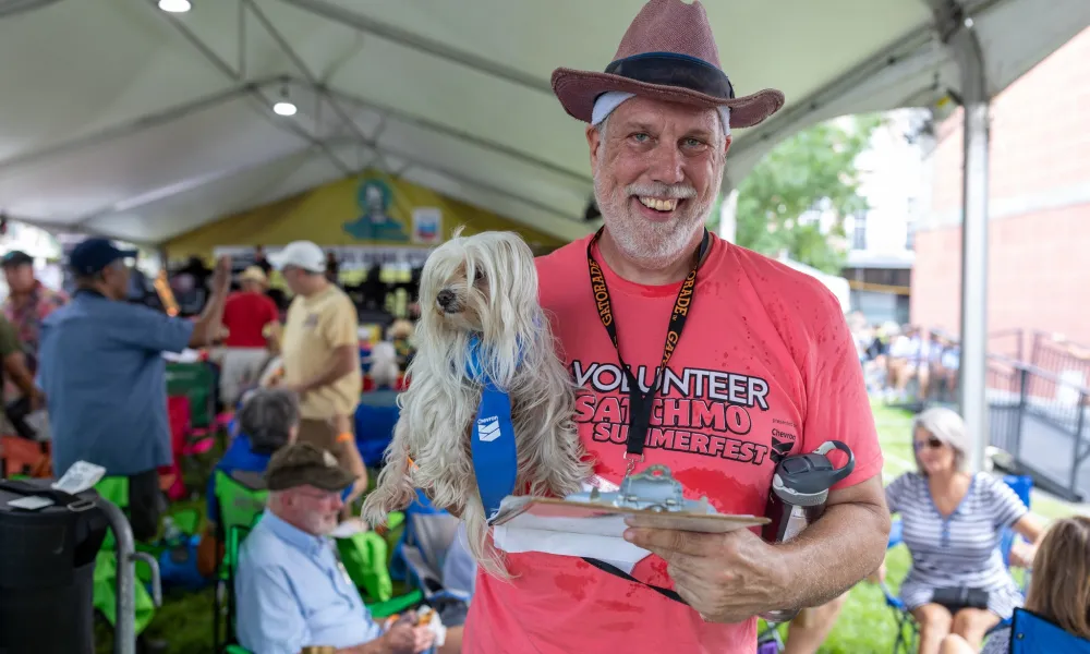 Smiling man in a pink volunteer shirt holding a small dog and clipboard at an outdoor festival tent.