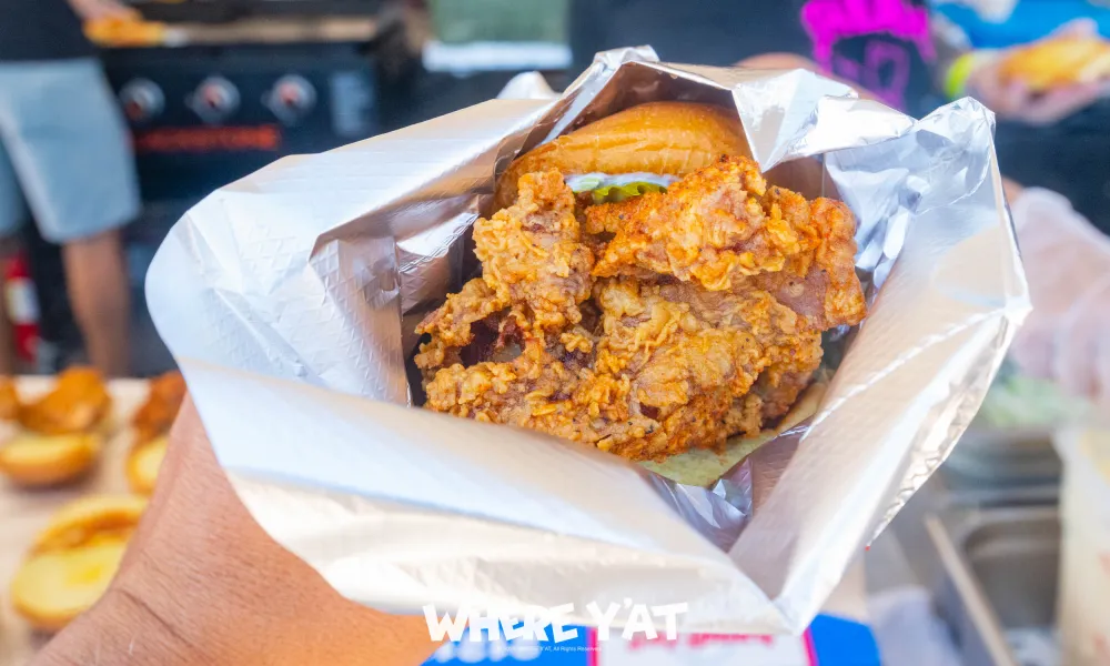 Hand holding a crispy fried chicken sandwich wrapped in foil with a bun and lettuce visible.