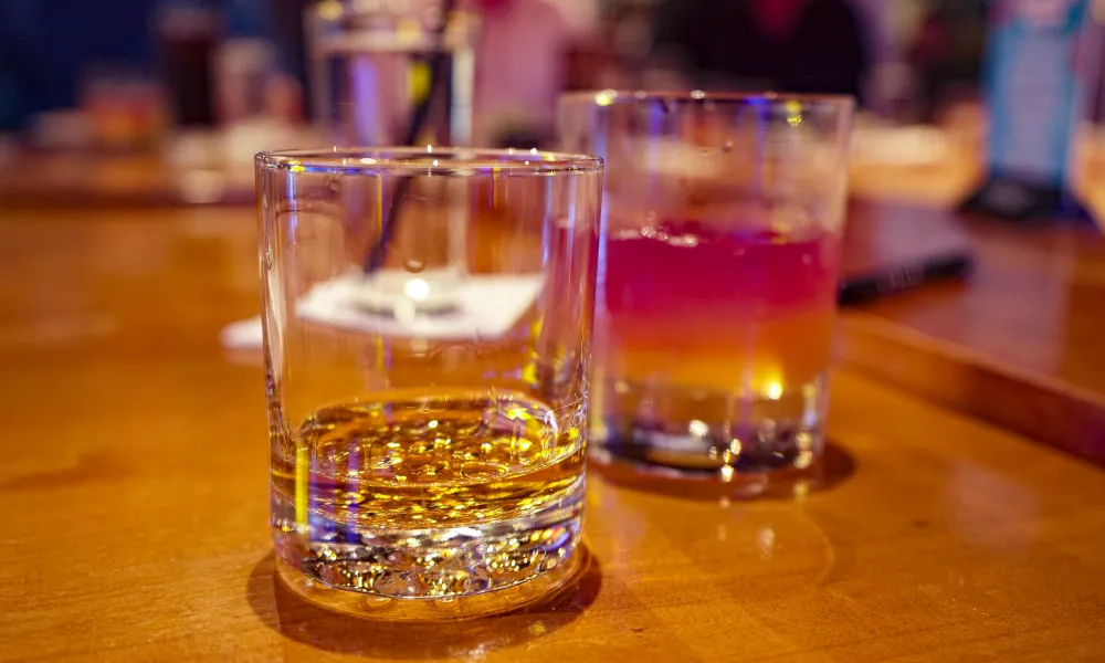 Two glasses on a wooden table, one with whiskey and the other with a layered cocktail, blurred background.