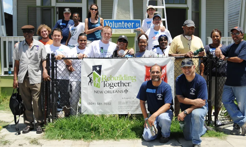 Group of diverse volunteers posing behind a Rebuilding Together New Orleans banner on a sunny day
