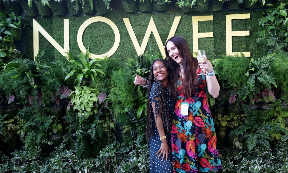 Two women smiling and posing in front of lush greenery with large NOWFE sign at an outdoor event.