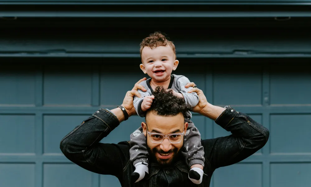 Smiling baby sitting on father's shoulders in front of a blue garage door on a casual day
