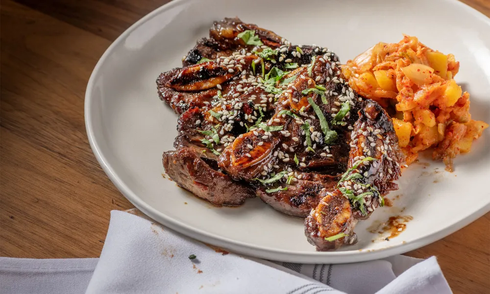 Grilled beef steak garnished with sesame seeds and herbs served with kimchi on a white plate.