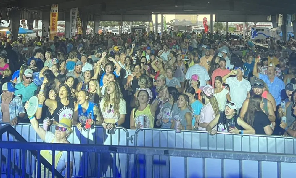 Large crowd enjoying an indoor concert or event with people cheering and taking photos under overhead lighting.