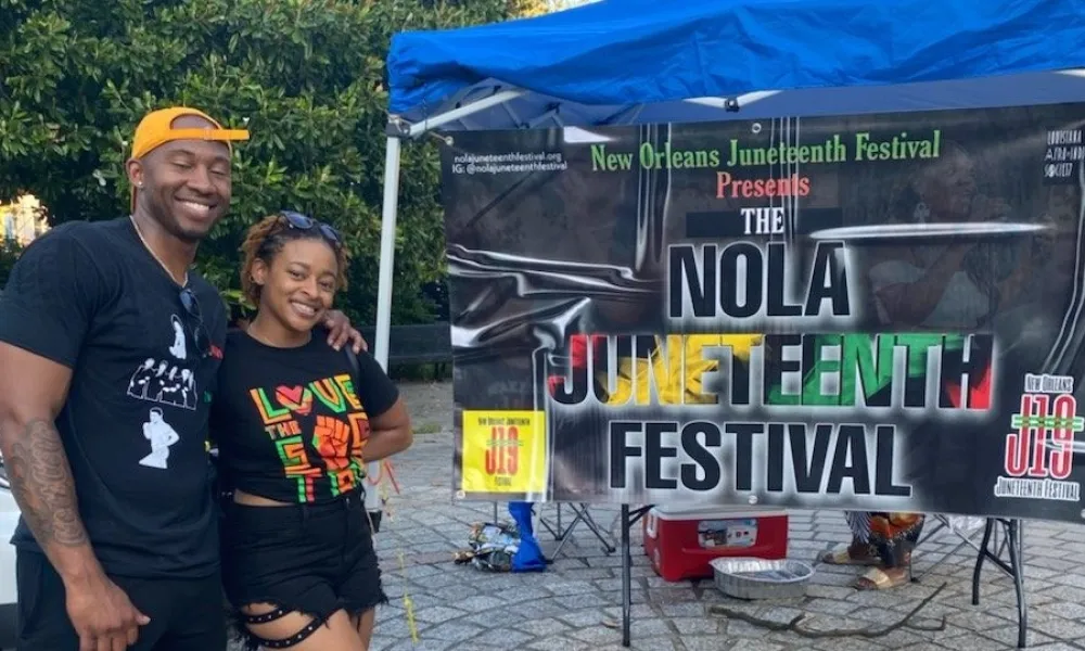 Smiling couple posing in front of a colorful NOLA Juneteenth Festival banner under a blue canopy on a cobblestone path.