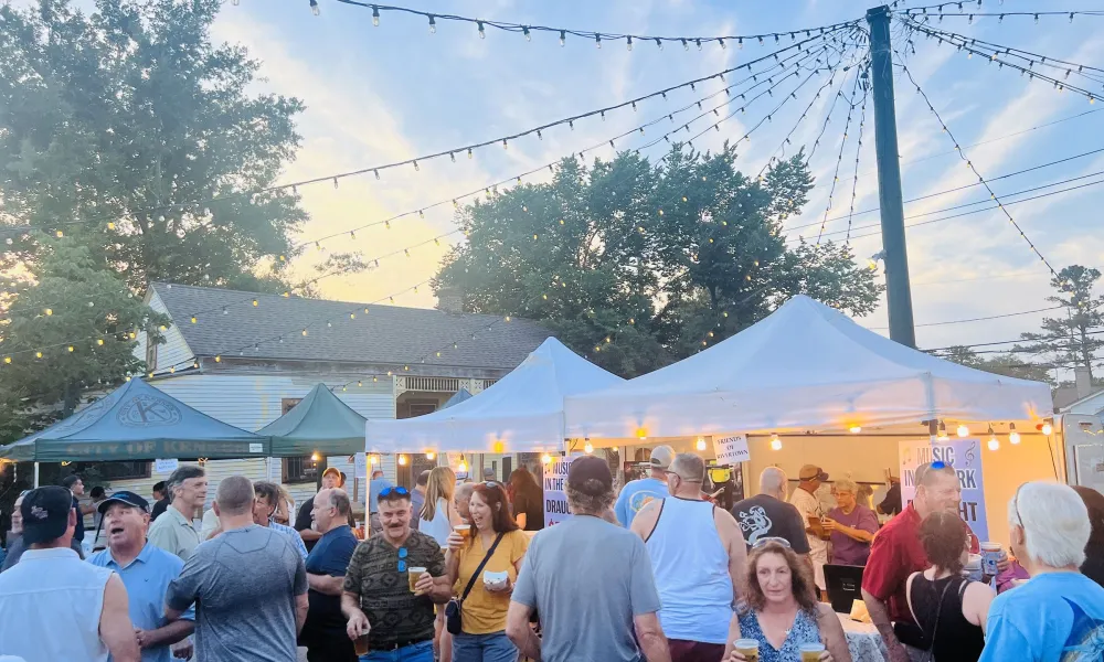 Crowd enjoying an outdoor festival with tents and string lights during early evening under a blue sky.
