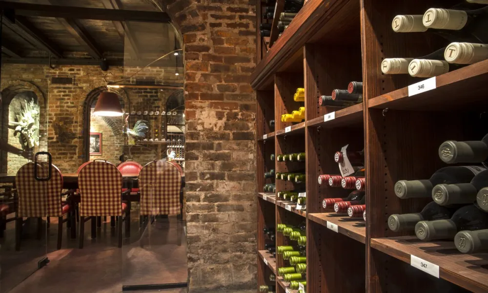 Wine bottles on wooden shelves in a dimly lit cellar dining room with exposed brick walls and checkered chairs.