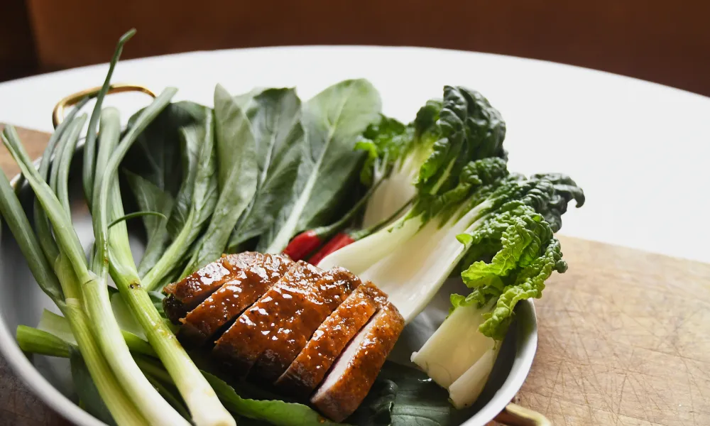 Plate with glazed sliced meat and fresh green vegetables including bok choy and scallions on wooden table.