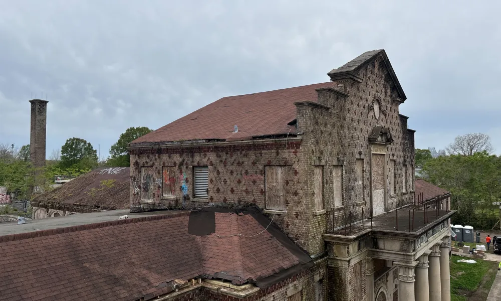 Abandoned brick building with boarded windows, red roof, and classical columns under a cloudy sky.