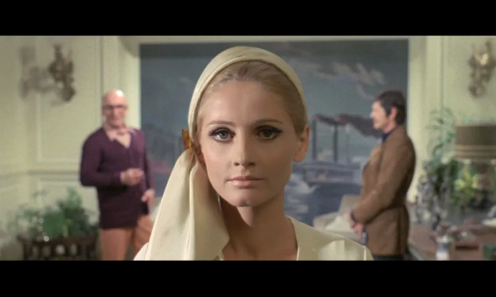 Close-up of a woman with makeup and headscarf, two men blurred in background inside a decorated room.