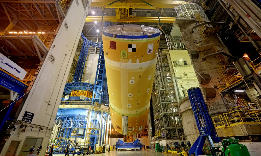 Large orange spacecraft fuel tank suspended inside a spacious industrial assembly facility with workers and machinery.