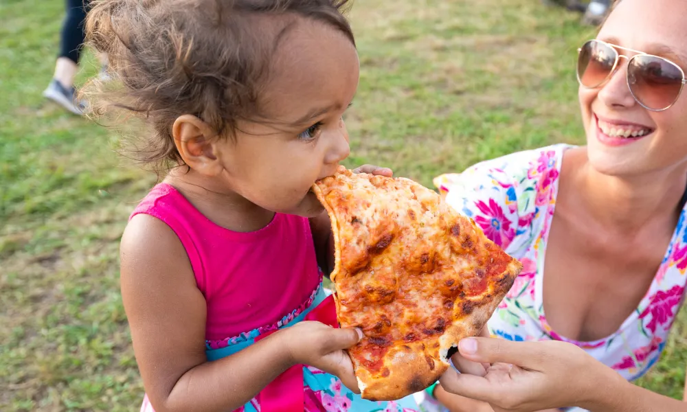 Young child wearing pink dress eating a large slice of pizza outdoors held by a smiling woman in sunglasses.
