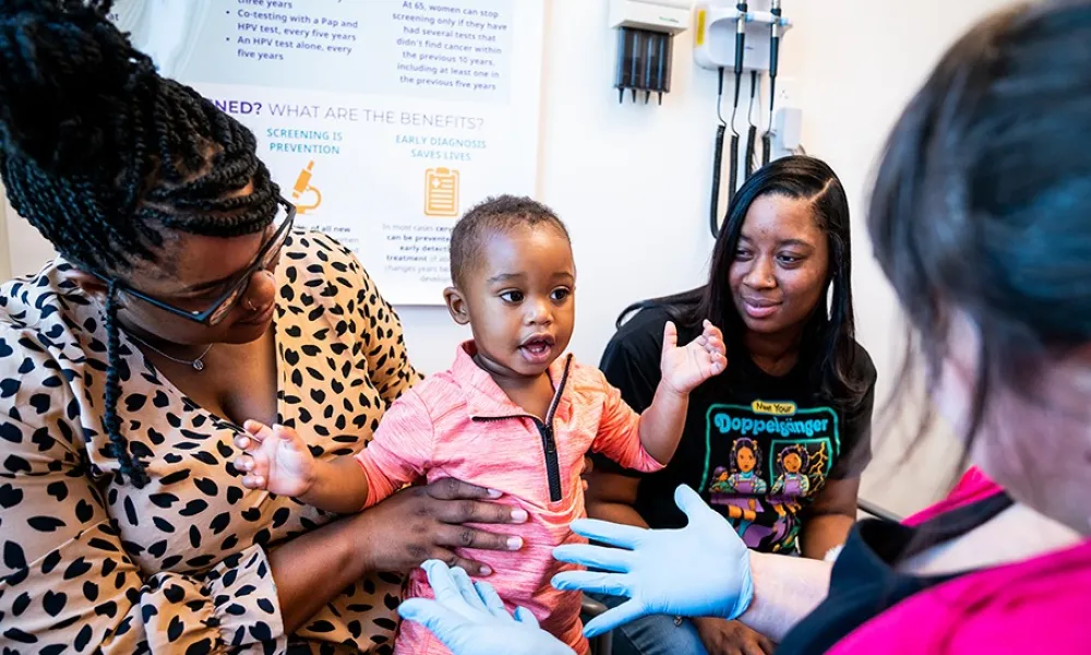 Child in pink shirt interacting with healthcare worker wearing gloves, accompanied by two women in a medical office.