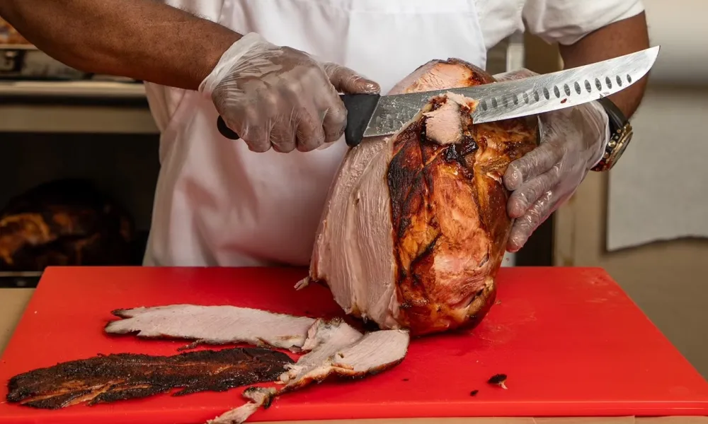 Chef slicing a large smoked ham on a red cutting board wearing gloves and white apron.