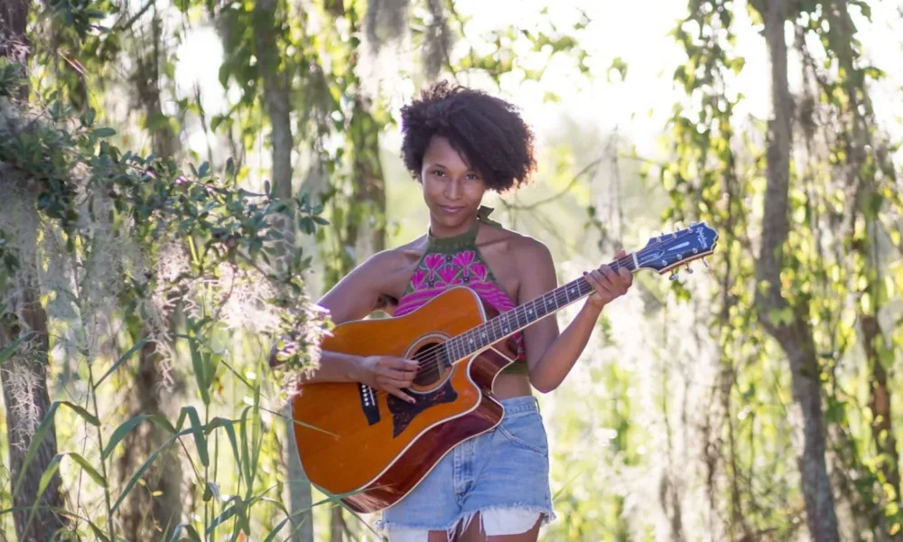 Young woman playing acoustic guitar barefoot in a sunny, lush forest clearing with hanging moss and sand.