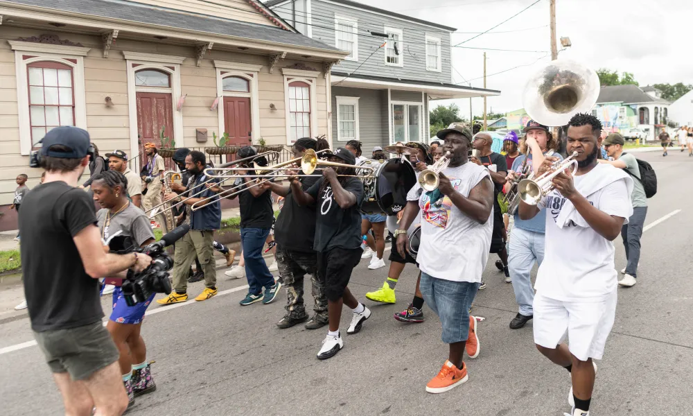 Street parade with musicians playing brass instruments and a cameraman filming the lively event.