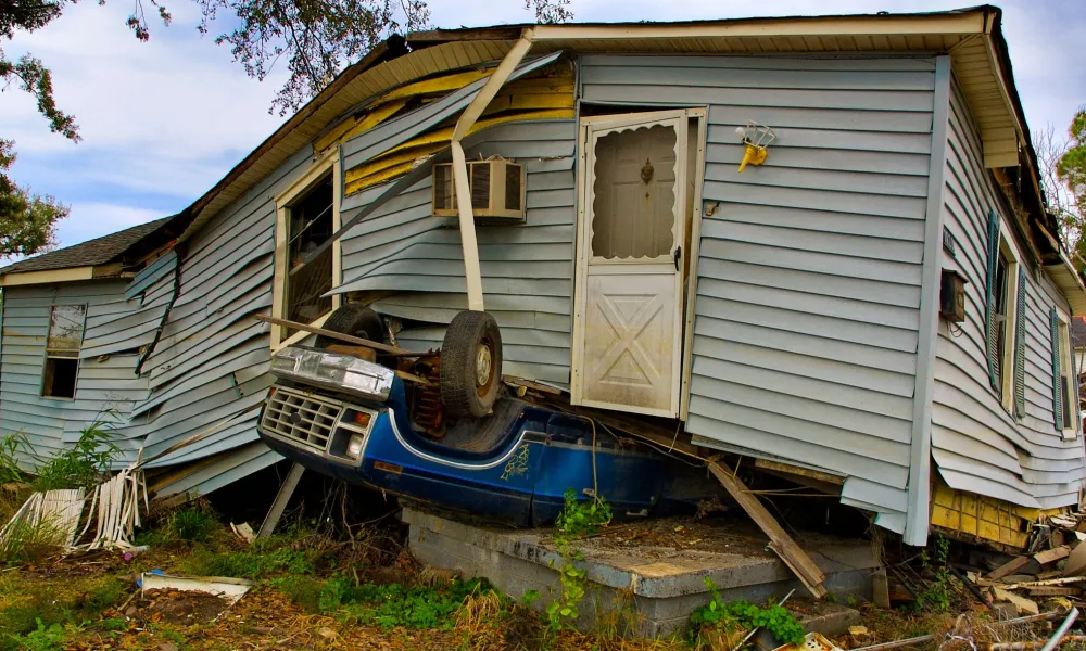 A damaged house with the front lifted by a blue car crushing the foundation amid debris and overgrown grass.