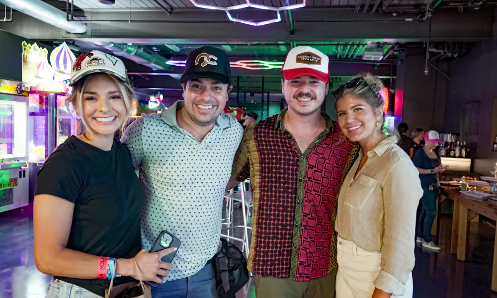 Four friends smiling in a vibrant arcade with colorful neon lights and casual attire including hats and wristbands.
