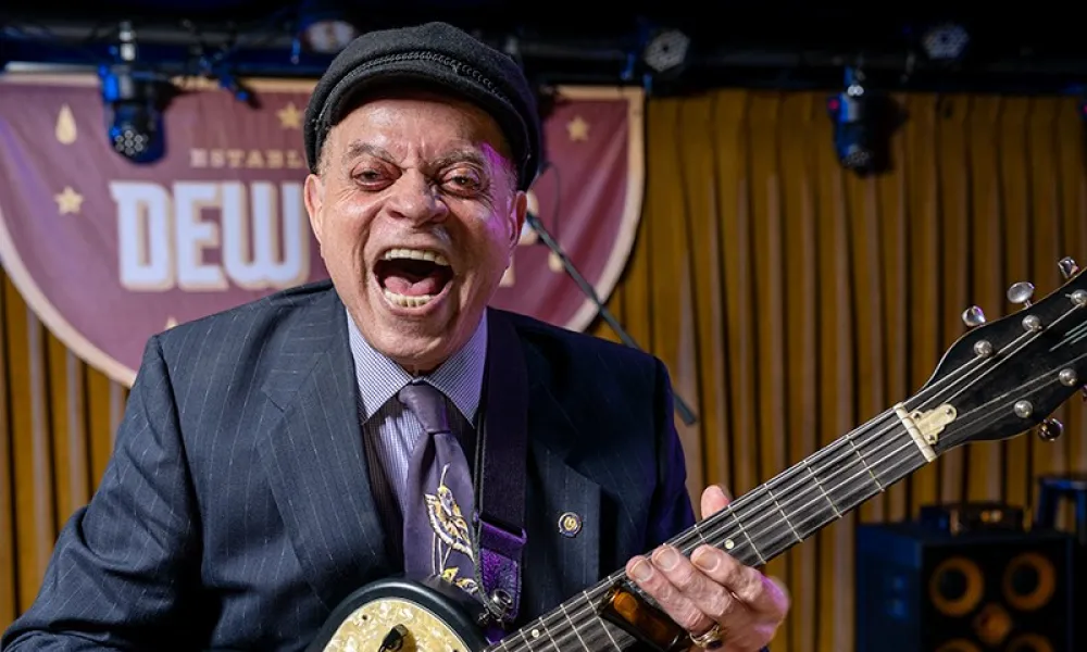 Smiling man in a suit and hat playing electric guitar on a stage with wooden backdrop and lights