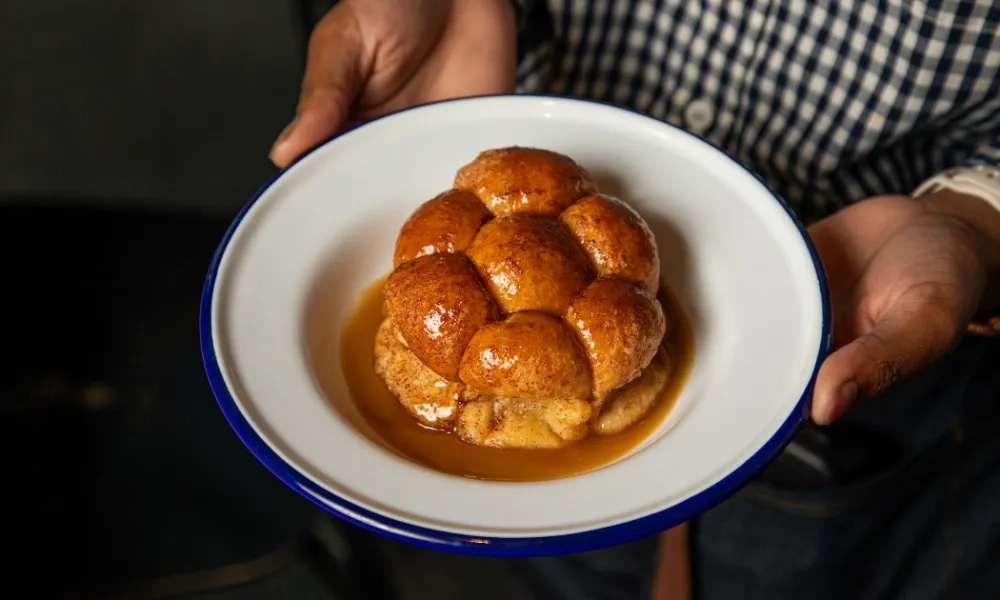Person holding a white plate with golden brown sticky buns drizzled in syrup inside a rustic setting