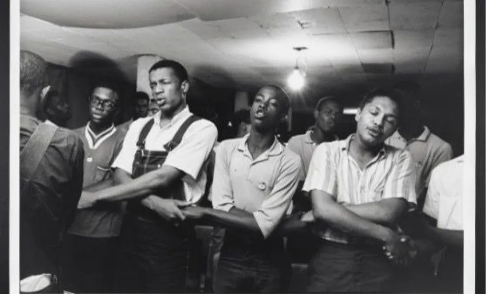 Black and white photo of young men standing with arms linked in a room with exposed ceiling light.