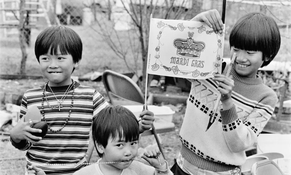 Three children smiling and playing outdoors, one holding a Mardi Gras flag, others wearing necklaces and festive clothes.