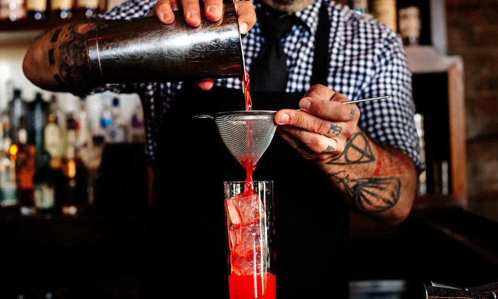 Bartender with tattoos pouring red cocktail through strainer into tall glass filled with ice at bar