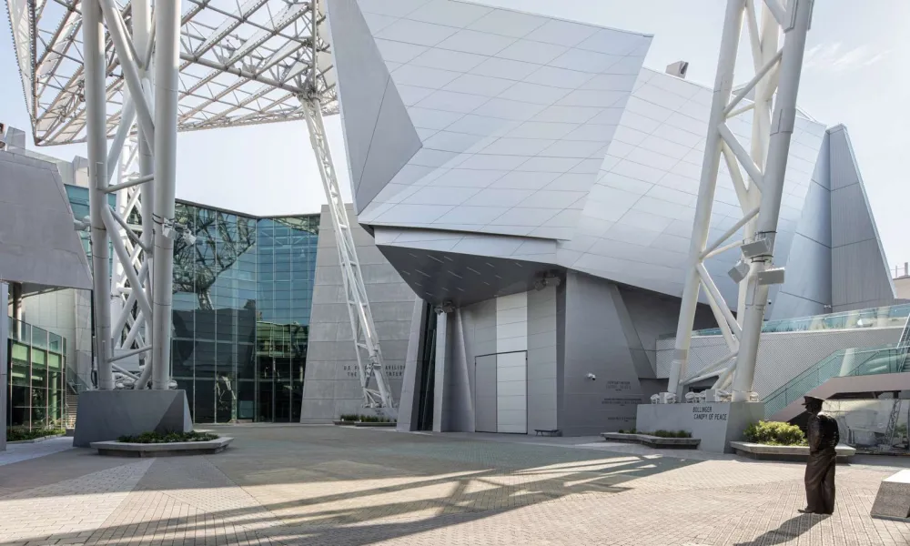 Modern architectural building with angular metal panels and large steel supports casting shadows on a paved courtyard.