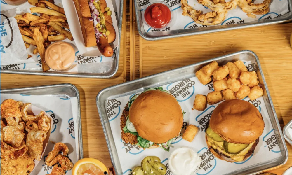 Assorted trays of fast food including burgers, hot dog, fries, tater tots, and fried snacks on wooden table