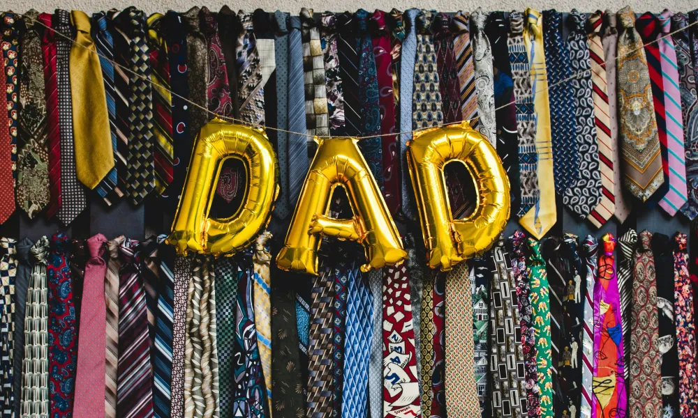 Gold balloons spelling DAD hang among rows of colorful patterned men's neckties on a wall display.