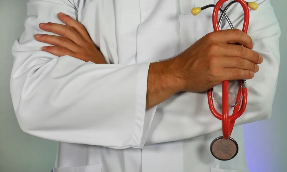 Doctor in white coat holding red stethoscope with arms crossed against light background