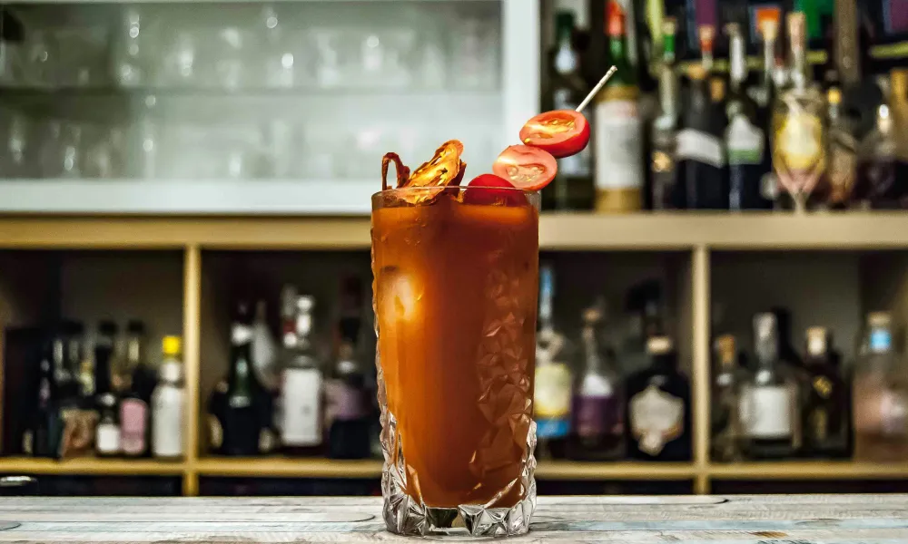 Tall Bloody Mary cocktail garnished with cherry tomatoes and dried chili on bar counter with bottles in background