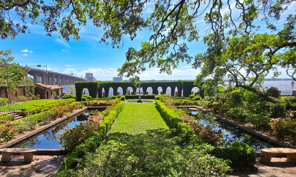 Elegant garden with manicured hedges, reflective pools, stone benches, and arched hedges overlooking a water body.