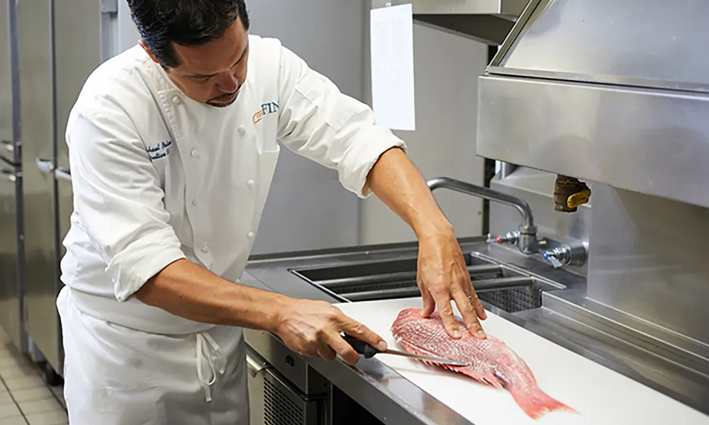 Chef filleting a fresh whole red fish on a stainless steel kitchen counter in a professional kitchen.