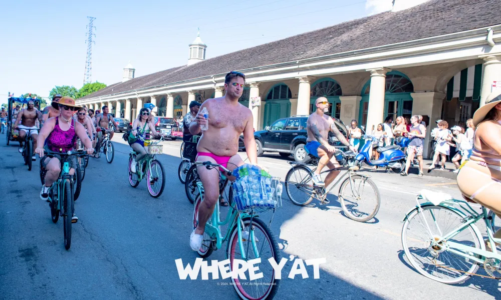 Group of minimally clothed cyclists riding on a sunny street during a festive event with spectators watching