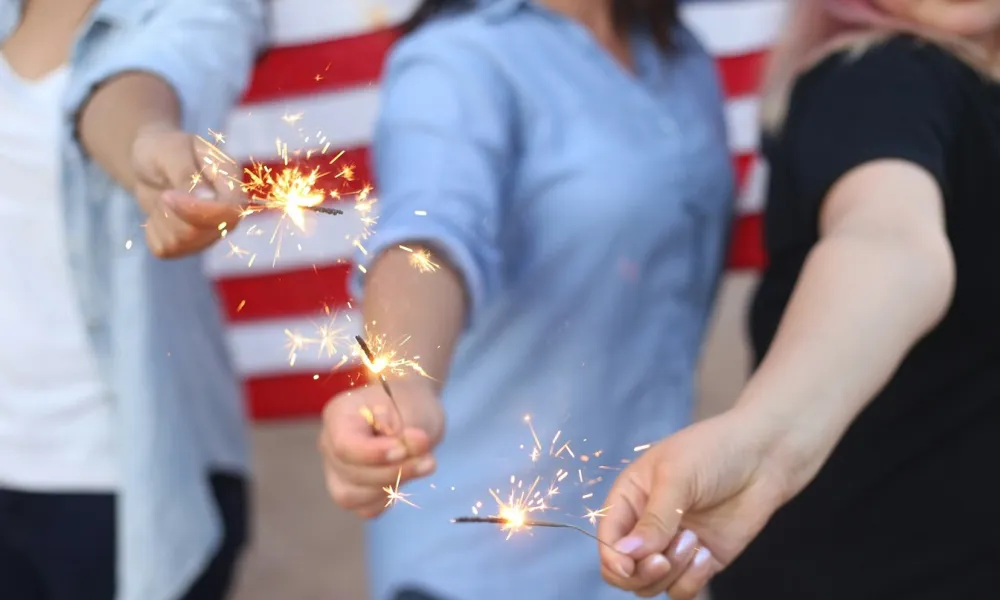 Three people holding lit sparklers with an American flag in the background celebrating a patriotic event