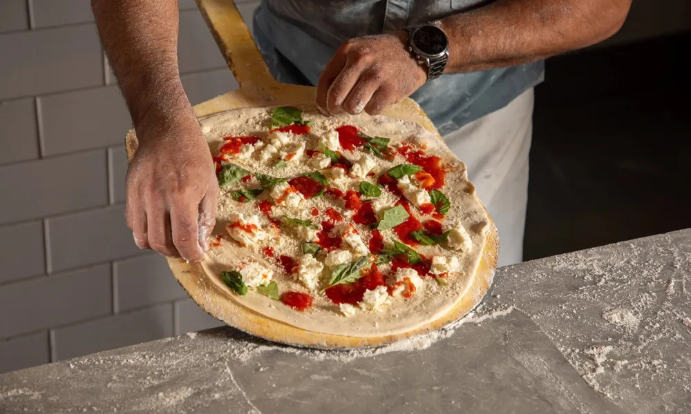 Person preparing homemade pizza with tomato sauce, cheese, and basil on a wooden paddle.