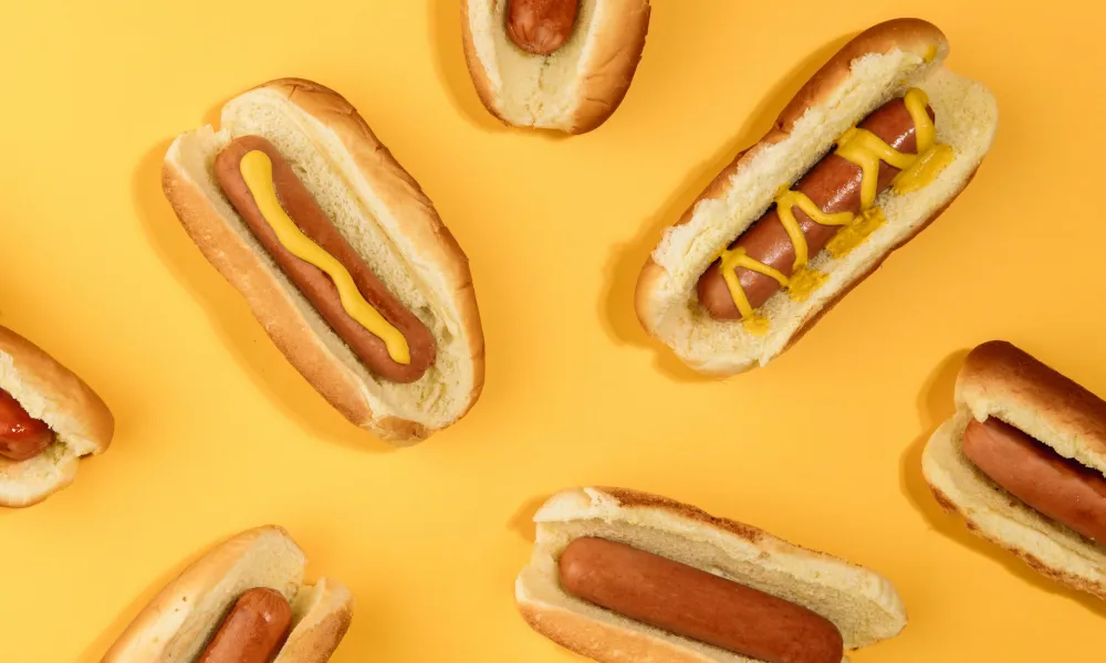 Several hot dogs in buns on a yellow background, some topped with yellow mustard in various patterns.