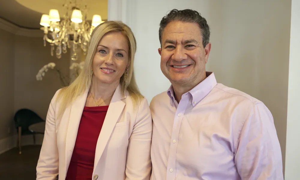 Smiling man and woman in business attire standing indoors with chandelier and elegant decor in the background
