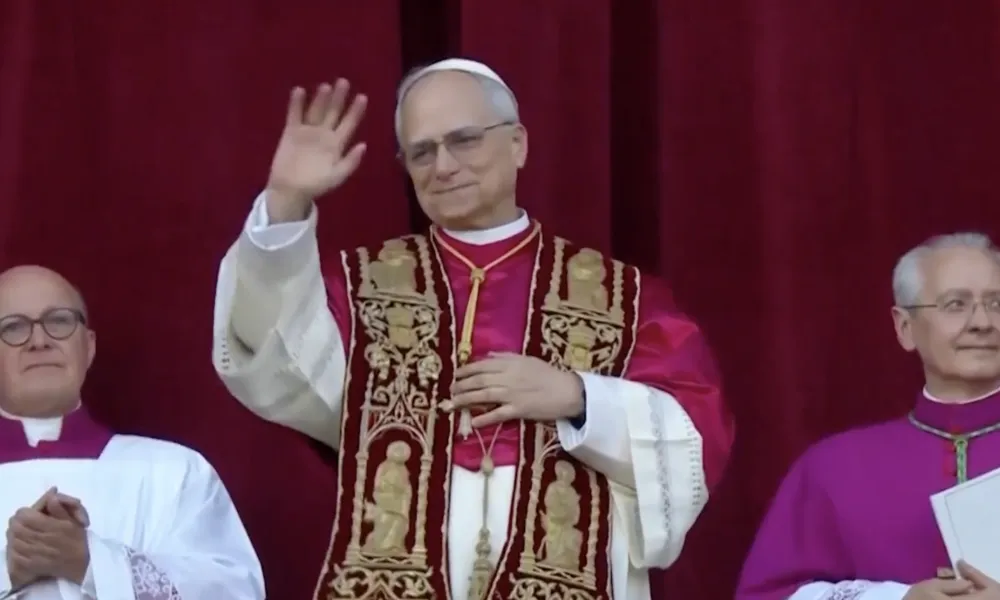 Pope in ornate vestments waving with two clergymen in ceremonial robes against red curtain background.