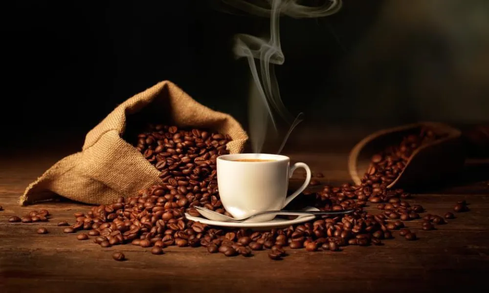 Steaming cup of coffee surrounded by spilled coffee beans from burlap sacks on a wooden surface.