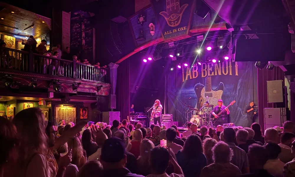Crowded indoor concert with musician performing on stage under purple lights and audience watching attentively
