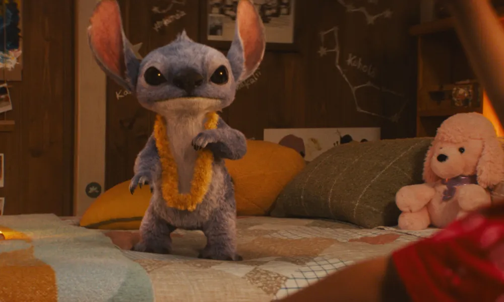 Blue alien creature wearing a yellow lei standing on a bed in a cozy room with photos and a stuffed dog toy.