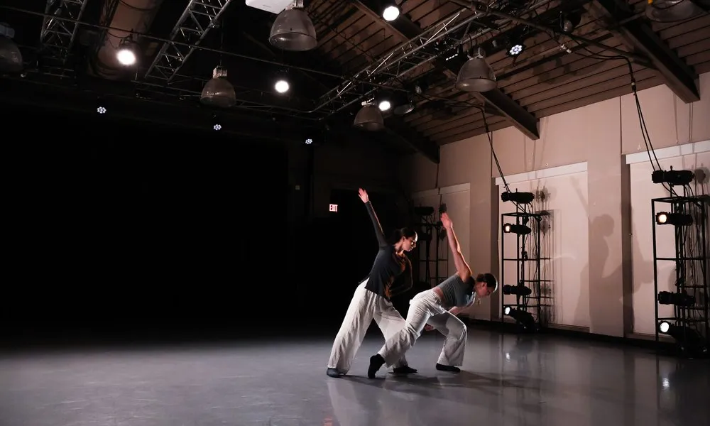 Two contemporary dancers performing in a dimly lit studio with spotlight and industrial ceiling beams.