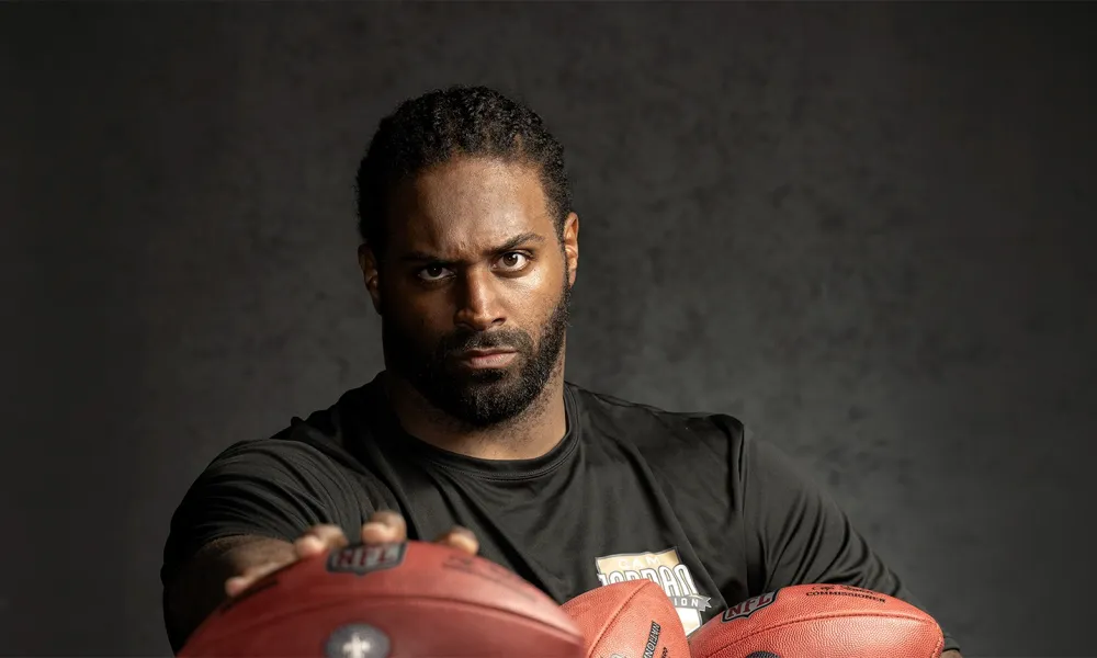 Serious male football player holding three footballs wearing a black shirt against a dark background