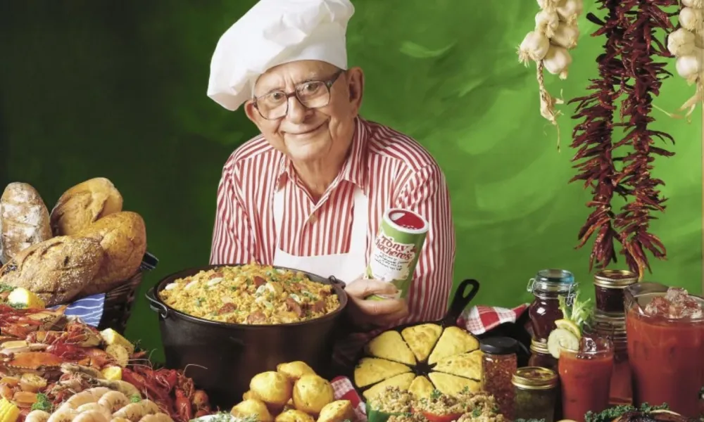 Elderly chef in white hat and red striped shirt presenting a large dish surrounded by bread, seafood, and vegetables.