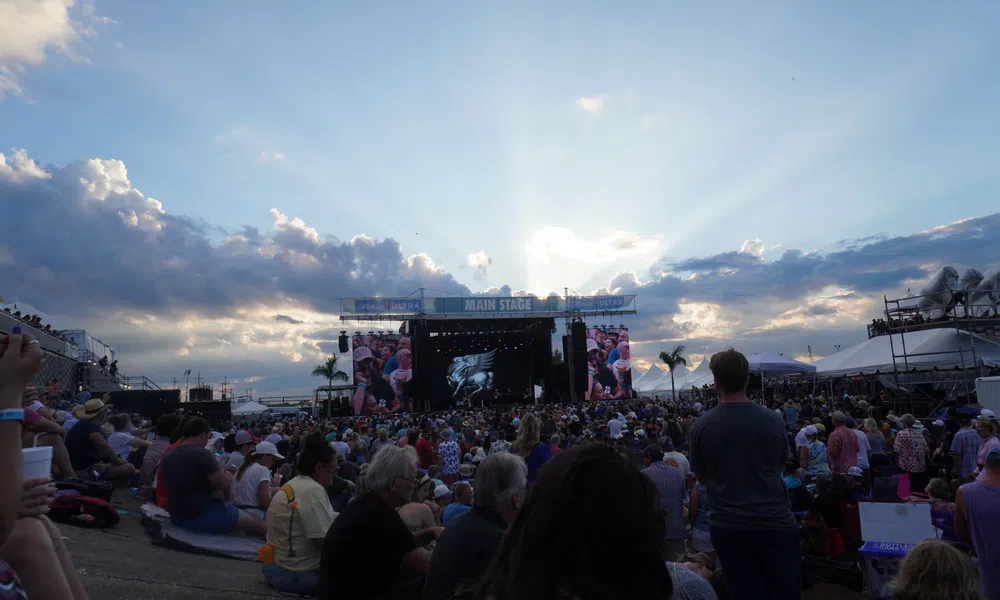 Huge crowd gathered at an outdoor music festival facing the main stage under a partly cloudy sky at sunset.