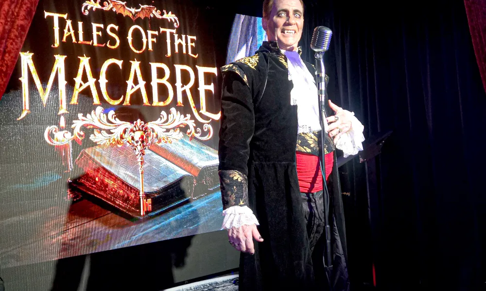 Performer in gothic costume stands next to a microphone with Tales of the Macabre sign behind him on stage.