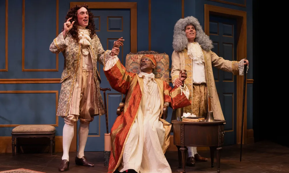 Three actors in 18th-century costumes performing on stage with blue paneled walls and period furniture.